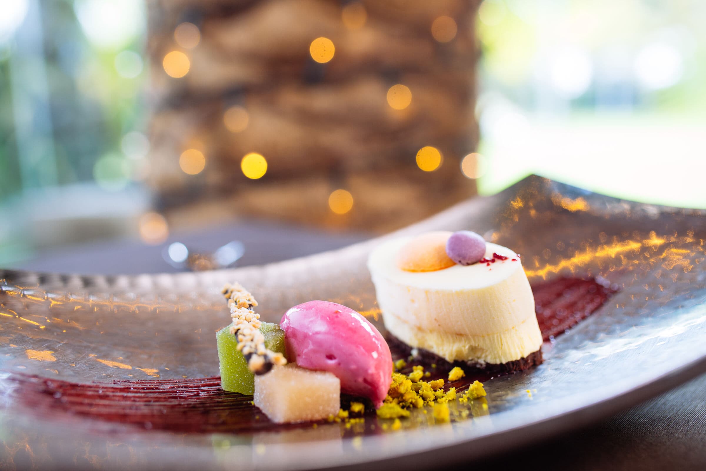 Postre de autor del catering de El Cachirulo sobre plato de metal martillado, con bokeh de guirnaldas cálidas