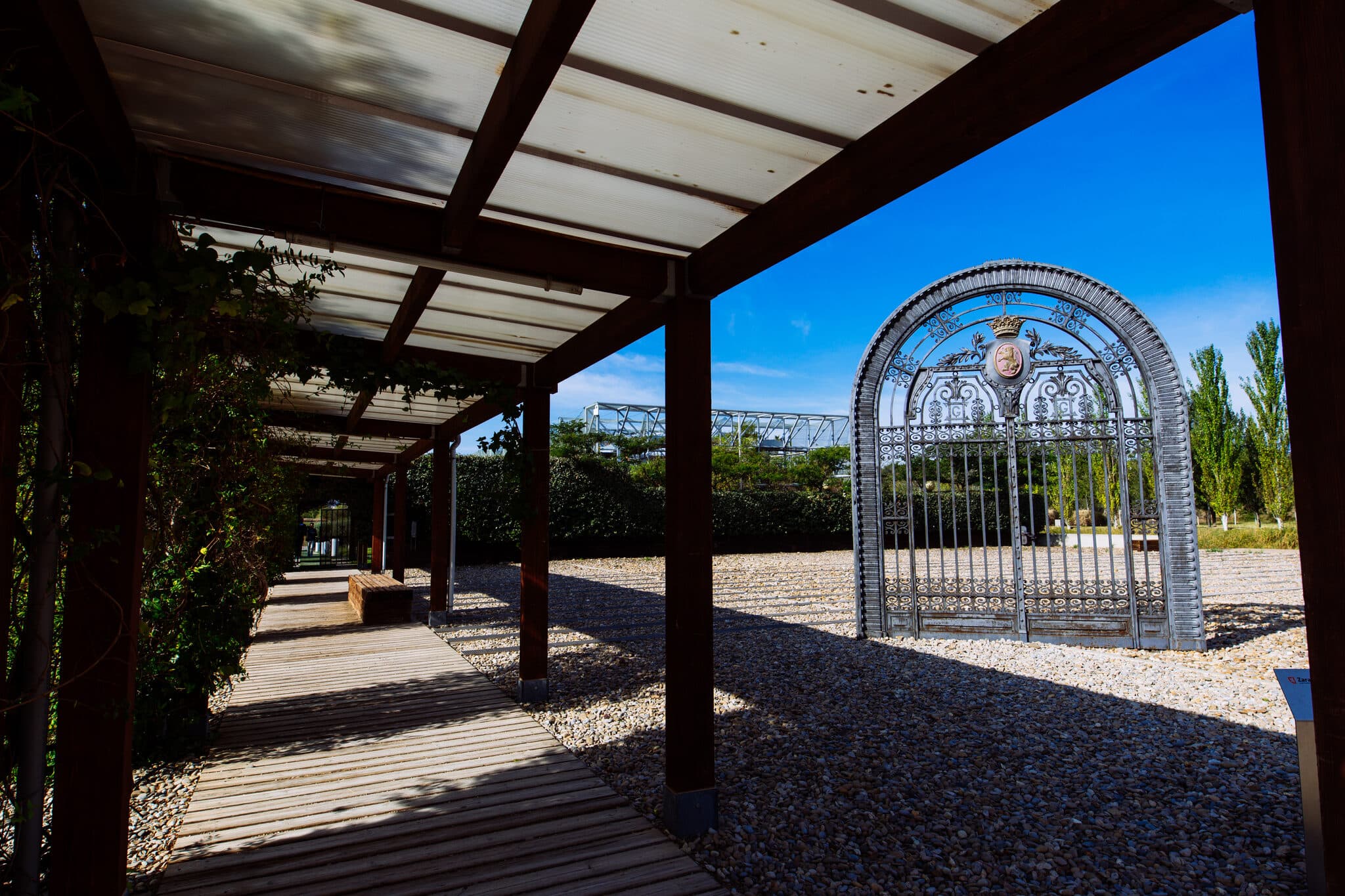La pérgola de madera del Pabellón de Ceremonias, abierta al arco de forja entre los jardines